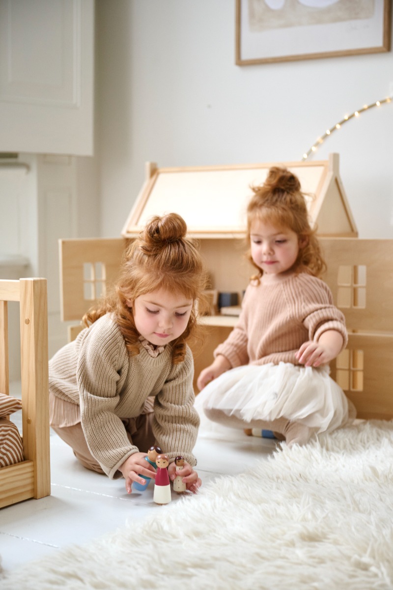 Deux enfants jouent avec des figurines en bois devant une maison de poupée en bois naturel, dans une chambre d’enfant lumineuse et chaleureuse.